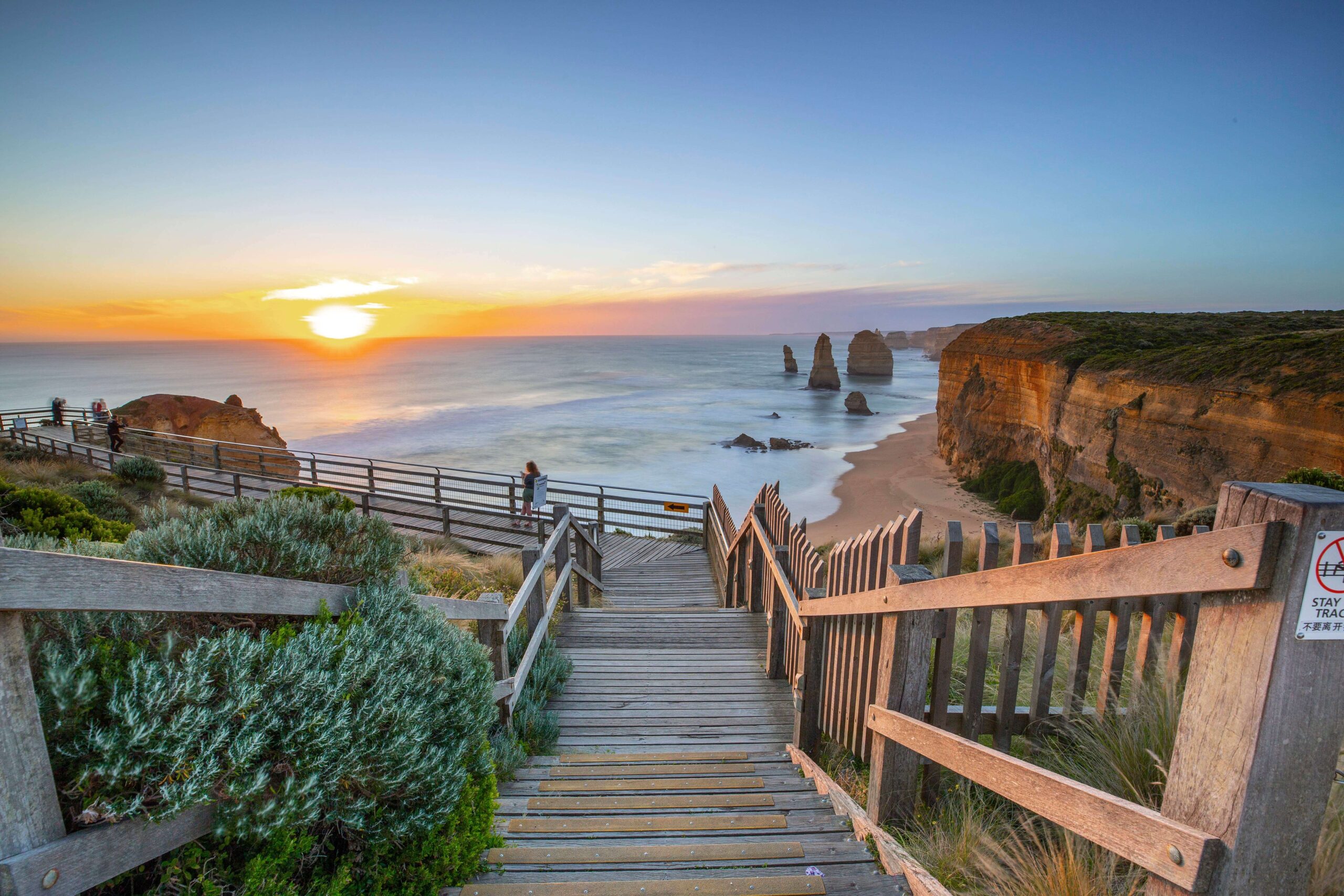Hoàng hôn tại Twelve Apostles trên Great Ocean Road, Úc với lối đi gỗ dẫn xuống bãi biển và các khối đá vôi ngoài khơi.