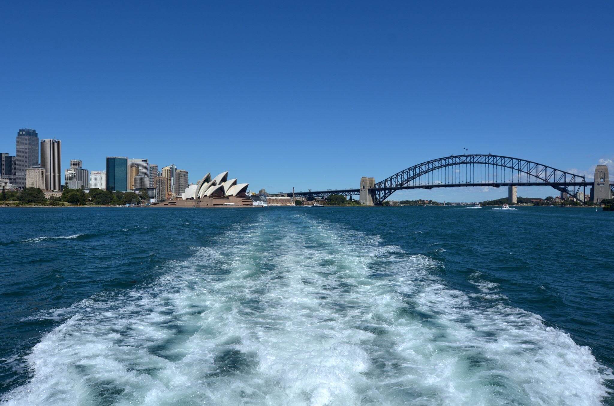 Sydney Harbour với Nhà hát Opera và cầu Harbour Bridge nhìn từ thuyền, mặt nước xanh và vệt sóng phía sau.
