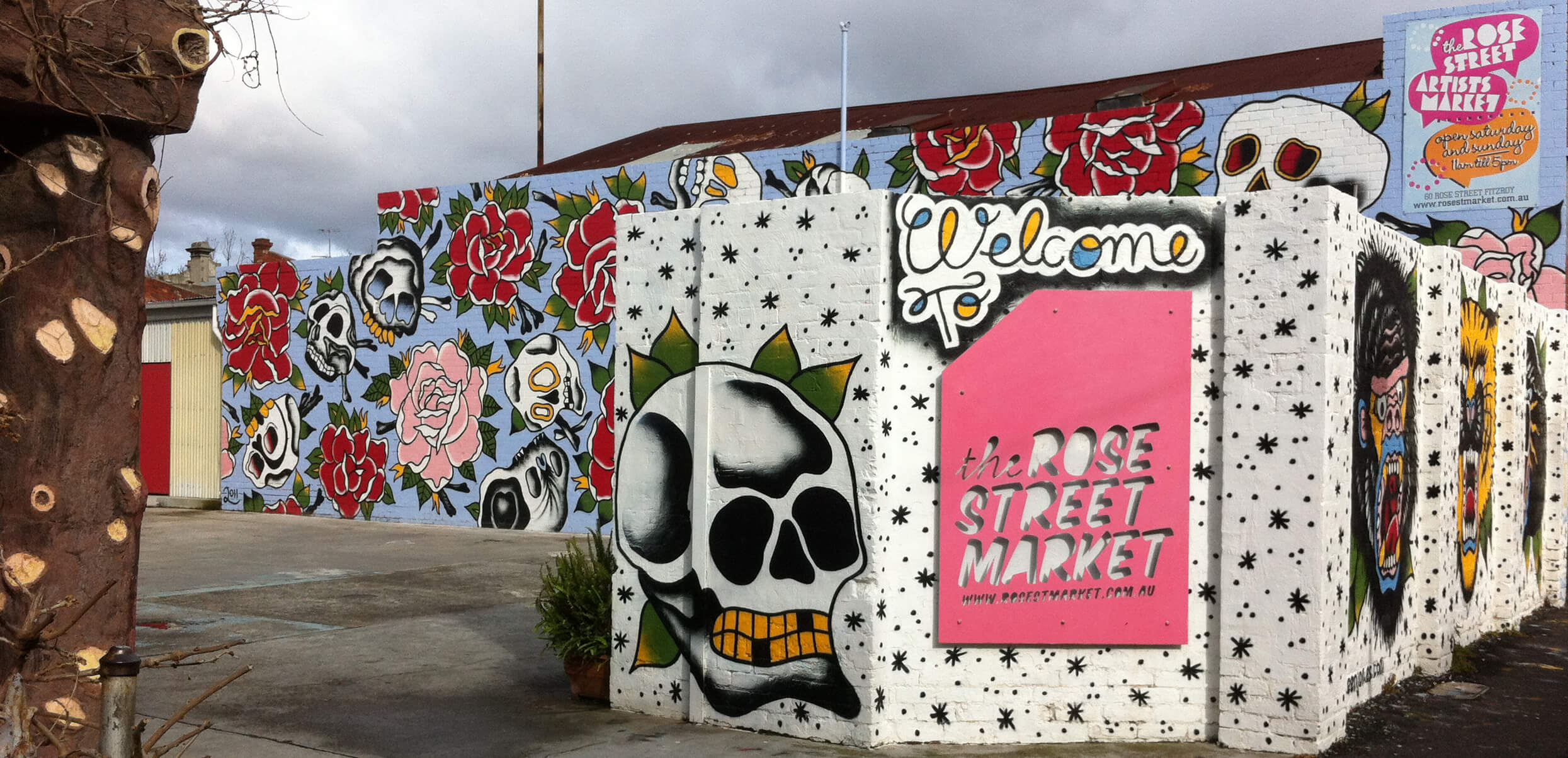 Bức tường đầy màu sắc tại The Rose Street Artists’ Market với các hình vẽ graffiti nổi bật như đầu lâu và hoa hồng. Biển hiệu “Welcome” và tên khu chợ tạo điểm nhấn cho không gian sáng tạo, phản ánh tinh thần nghệ thuật đường phố đặc trưng của khu vực Fitzroy.
