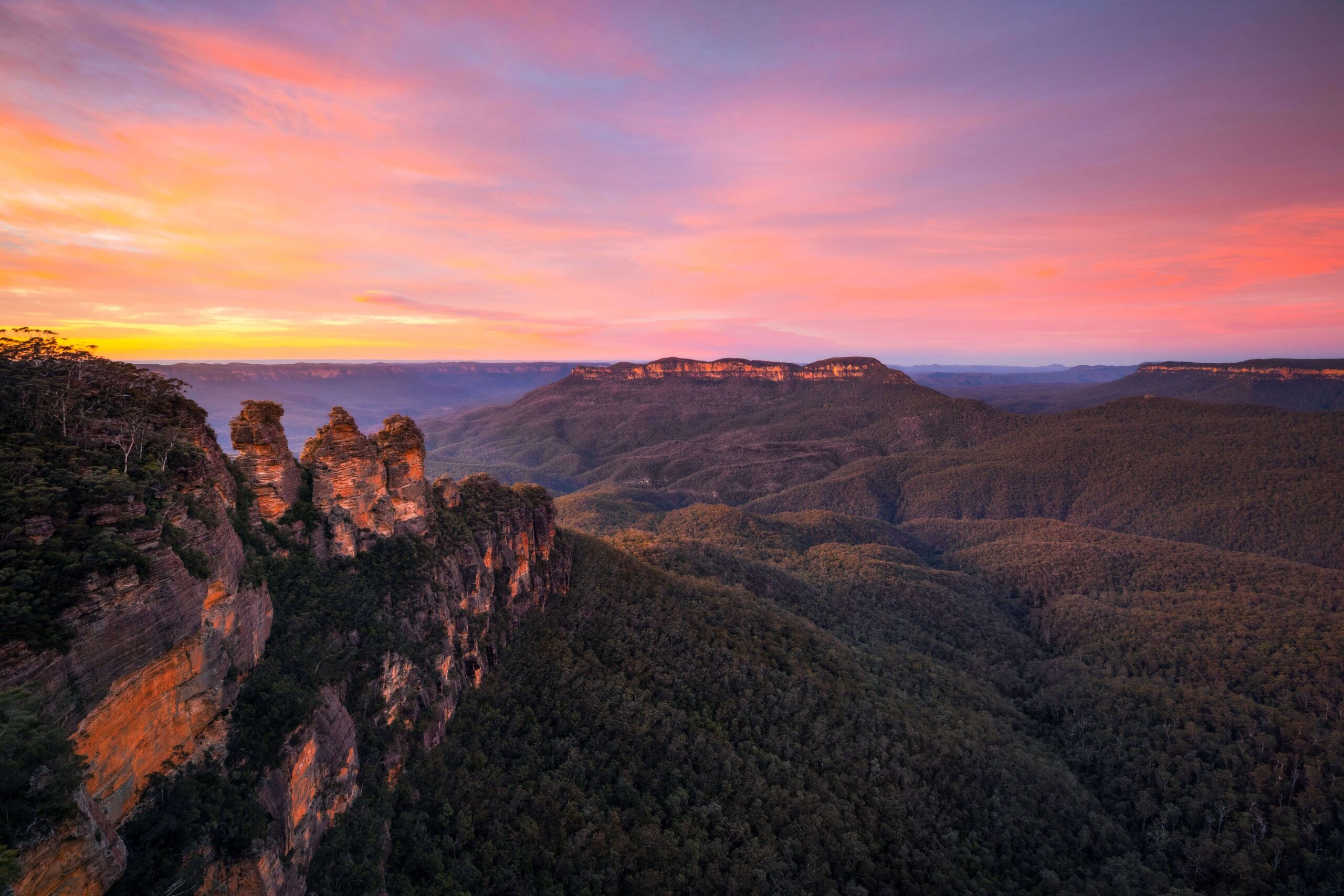 Khung cảnh Three Sisters tại Blue Mountains lúc hoàng hôn, nhìn ra thung lũng Jamison hùng vĩ với những vách đá sa thạch đặc trưng – một biểu tượng thiên nhiên nổi tiếng của Úc.