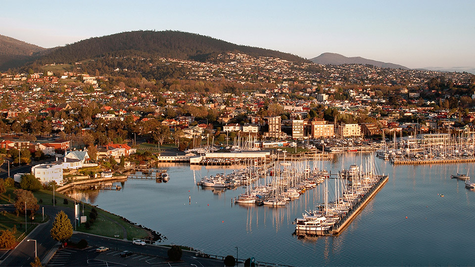 Toàn cảnh thành phố Hobart, Tasmania với bến cảng nhộn nhịp, những chiếc thuyền neo đậu và khu dân cư trải dài dưới chân núi, tạo nên khung cảnh yên bình và đặc trưng của thành phố cảng lâu đời tại Úc.