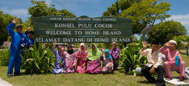 Nhóm người dân địa phương tại Home Island, Cocos (Keeling) Islands, Australia chào đón du khách trước bảng hiệu “Welcome to Home Island”, thể hiện nét văn hóa thân thiện và đời sống cộng đồng đặc trưng của đảo.