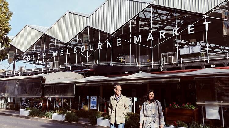 Hình ảnh bên ngoài South Melbourne Market với kiến trúc mái chợ đặc trưng và dòng chữ “South Melbourne Market” nổi bật, phía trước có hai người đang dạo bước, thể hiện không khí nhộn nhịp và trải nghiệm đời sống địa phương tại Melbourne.