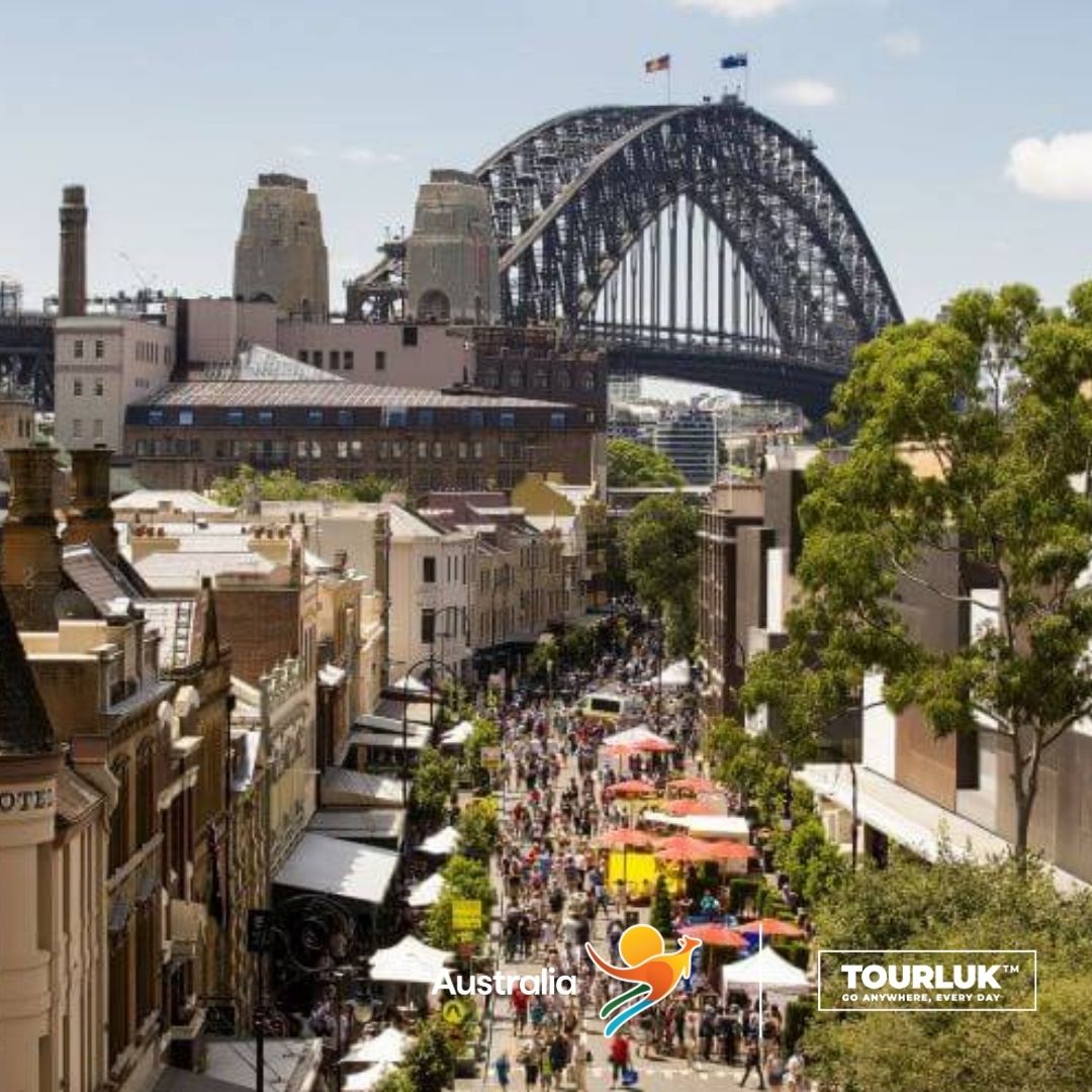 Khung cảnh nhộn nhịp tại khu phố cổ The Rocks ở Sydney, Australia với đông du khách tham quan, các gian hàng ngoài trời và cầu cảng Sydney Harbour Bridge nổi bật phía sau vào ban ngày.