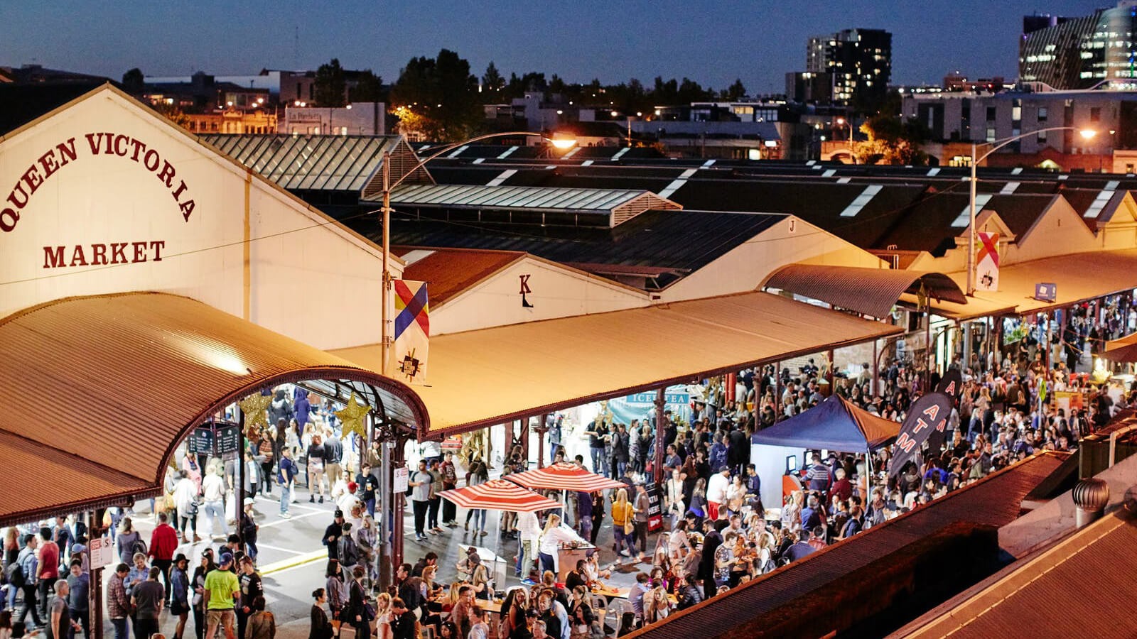 Khung cảnh nhộn nhịp tại Queen Victoria Market vào buổi tối, với đông đảo người dân và du khách tham quan, mua sắm và thưởng thức ẩm thực đường phố. Các gian hàng sáng đèn, mái che đặc trưng của khu chợ tạo nên không khí sôi động giữa lòng thành phố Melbourne.